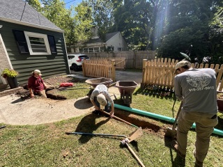 Crew preparing a drainage trench beside an existing sidewalk with green solid pipe ready for installation
