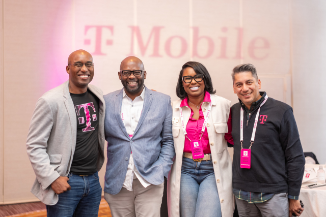 Four attendees posing together in front of the T-Mobile logo and magenta event lighting.