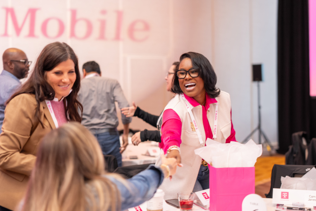 Attendees smiling and greeting one another around a table beneath the projected T-Mobile logo.